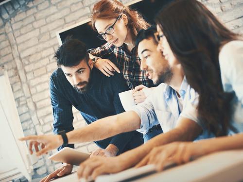 Closeup side view of group of IT experts completing a task in their office. There are two men and two women gathered around dual screen computer. One of the men is leading the project.